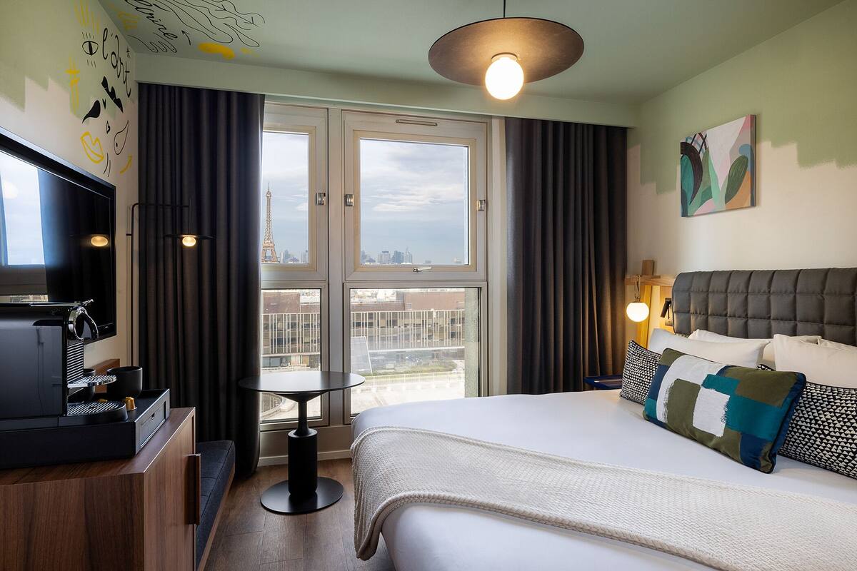 A hotel room with a bed, a TV, a coffee machine, a table, and a view of the Eiffel Tower.