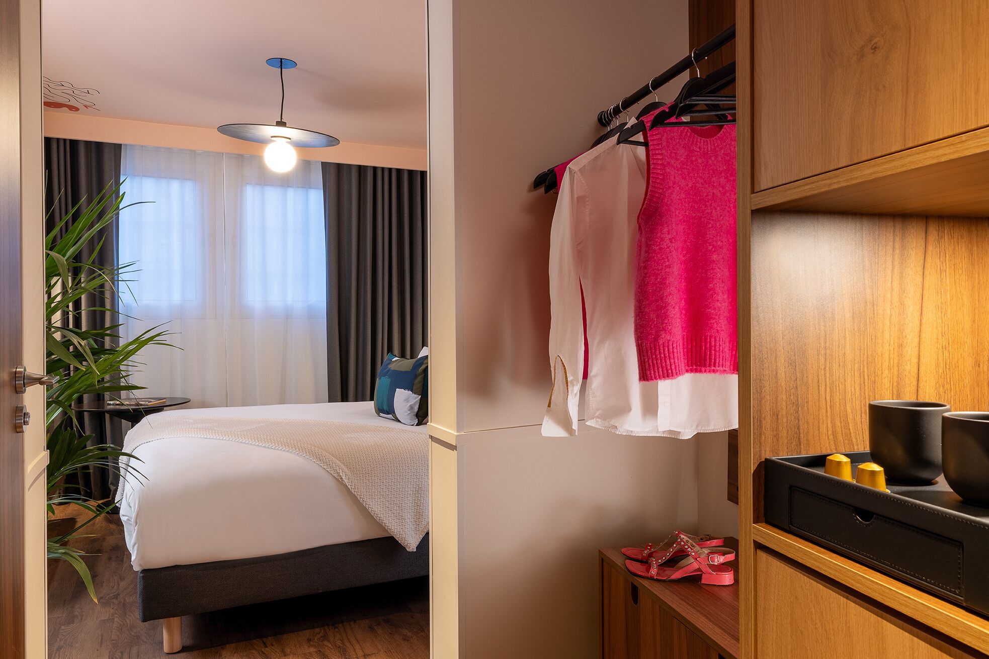 Quarto de hotel com cama, mesa de cabeceira, luminária pendente, guarda-roupa e sapateira.