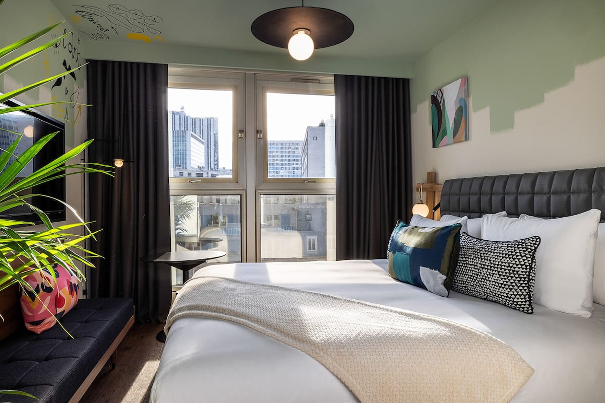 A modern hotel room with a large bed, a small table, a potted plant, and a view of the city through large windows.