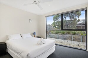A bedroom with a bed, a nightstand, a lamp, and a large window with a view of a garden and trees.