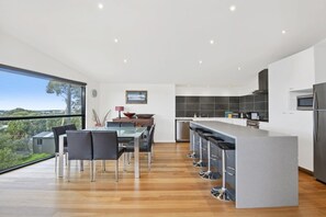 A modern kitchen with a bar area, a dining table with chairs, and a view through large windows.
