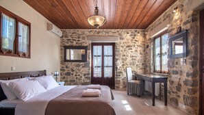 Habitació amb llit, sostre de fusta, parets de pedra, mirall i bany amb rentapla i cadira.