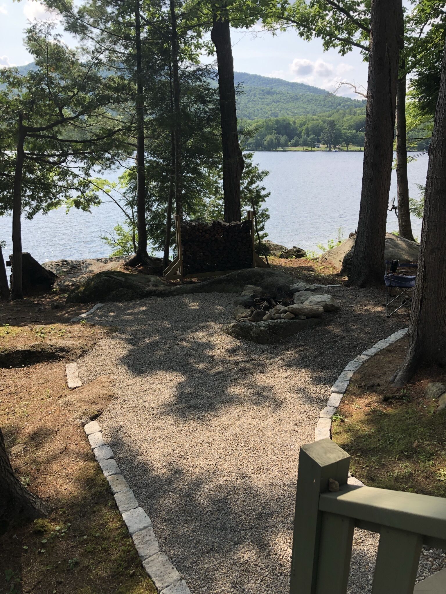 Un espace recouvert de gravier, avec un foyer en pierre, entouré d’arbres et offrant une vue sur un lac.