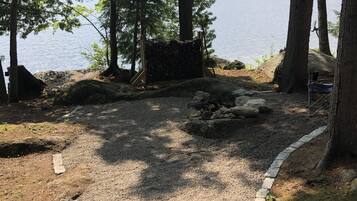 Un espace recouvert de gravier, avec un foyer en pierre, entouré d’arbres et offrant une vue sur un lac.