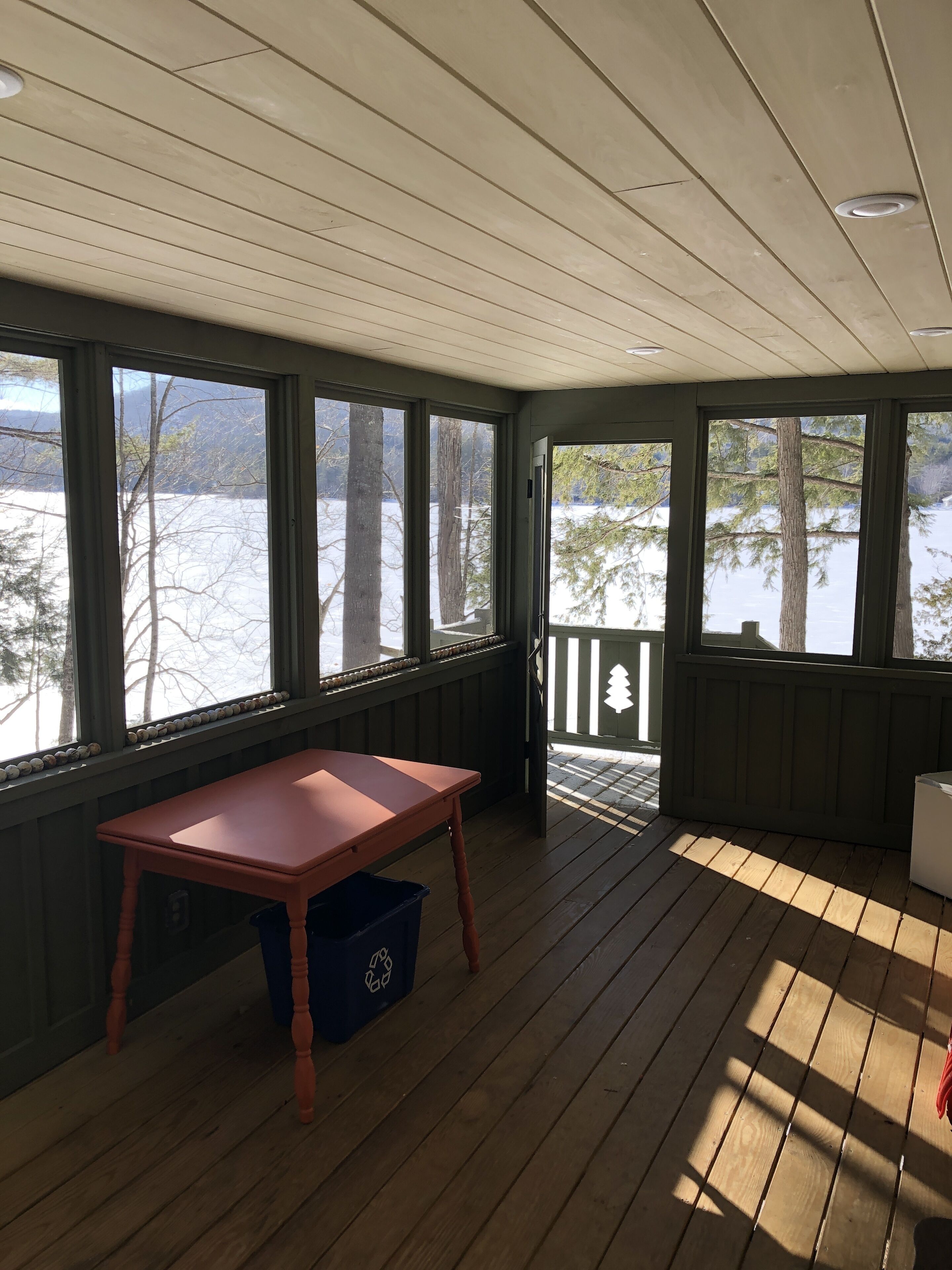 Une véranda couverte avec une table en bois et une poubelle, donnant sur un paysage enneigé parsemé d’arbres et surmonté d’un quai.