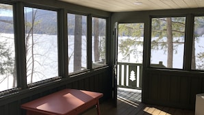 Varanda coberta com mesa de madeira e lixeira, com vista para uma paisagem nevada com árvores e um píer.