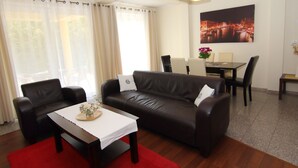 Una sala de estar con un sofá de cuero marrón, dos sillones, una mesa de centro con un jarrón de flores, una mesa de comedor con sillas, y un cuadro de un paisaje urbano en la pared.