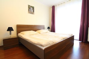 A neatly made bed in a room with wooden flooring, a nightstand with a lamp, and a painting on the wall.