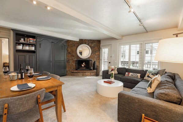 A living room with a fireplace, a dining area with a wooden table and chairs, and a gray sofa with decorative pillows. - Pool Breeze Condo steps from Vail MTN Gondola w/ fireplace, hot tub + spa access (Vail)
