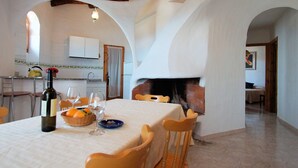 Une salle à manger avec une table dressée pour deux personnes, une bouteille de vin et un panier d’oranges. On aperçoit une cheminée au fond.
