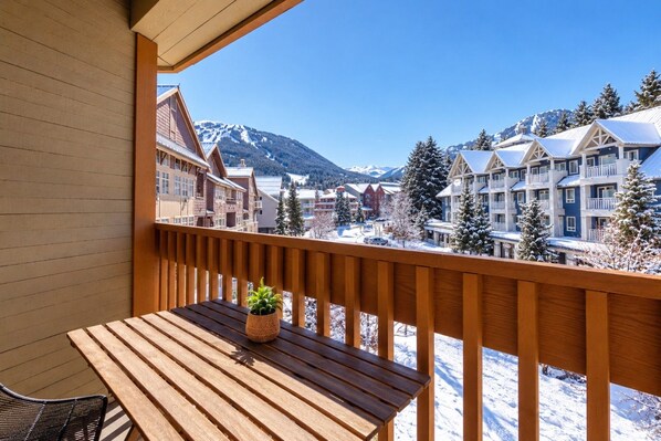 A wooden balcony with a table and a potted plant, overlooking a snowy mountain town with buildings and trees.