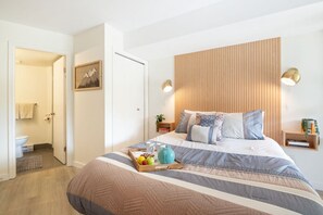 Quarto moderno com cabeceira de madeira, cama com roupa de cama listrada, mesas de cabeceira com abajures e uma bandeja de frutas sobre a cama.