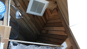 Una pequeña cabaña de madera con techo inclinado, un respiradero blanco, un perchero metálico y una puerta de madera con una toalla colgada.