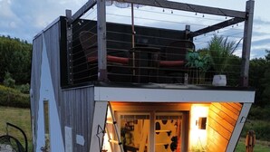 Una cabaña pequeña de dos pisos, con un balcón y una escalera que lleva a la entrada, rodeada de plantas en macetas.