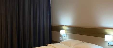Habitación de hotel con una cama grande, cortinas oscuras, cabecero con luces fijadas a la pared y suelo de madera.