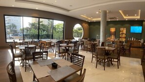 Un restaurante amplio con varias mesas y sillas, ventanas grandes que ofrecen vistas al exterior, un pilar central y una zona de recepción con un televisor.