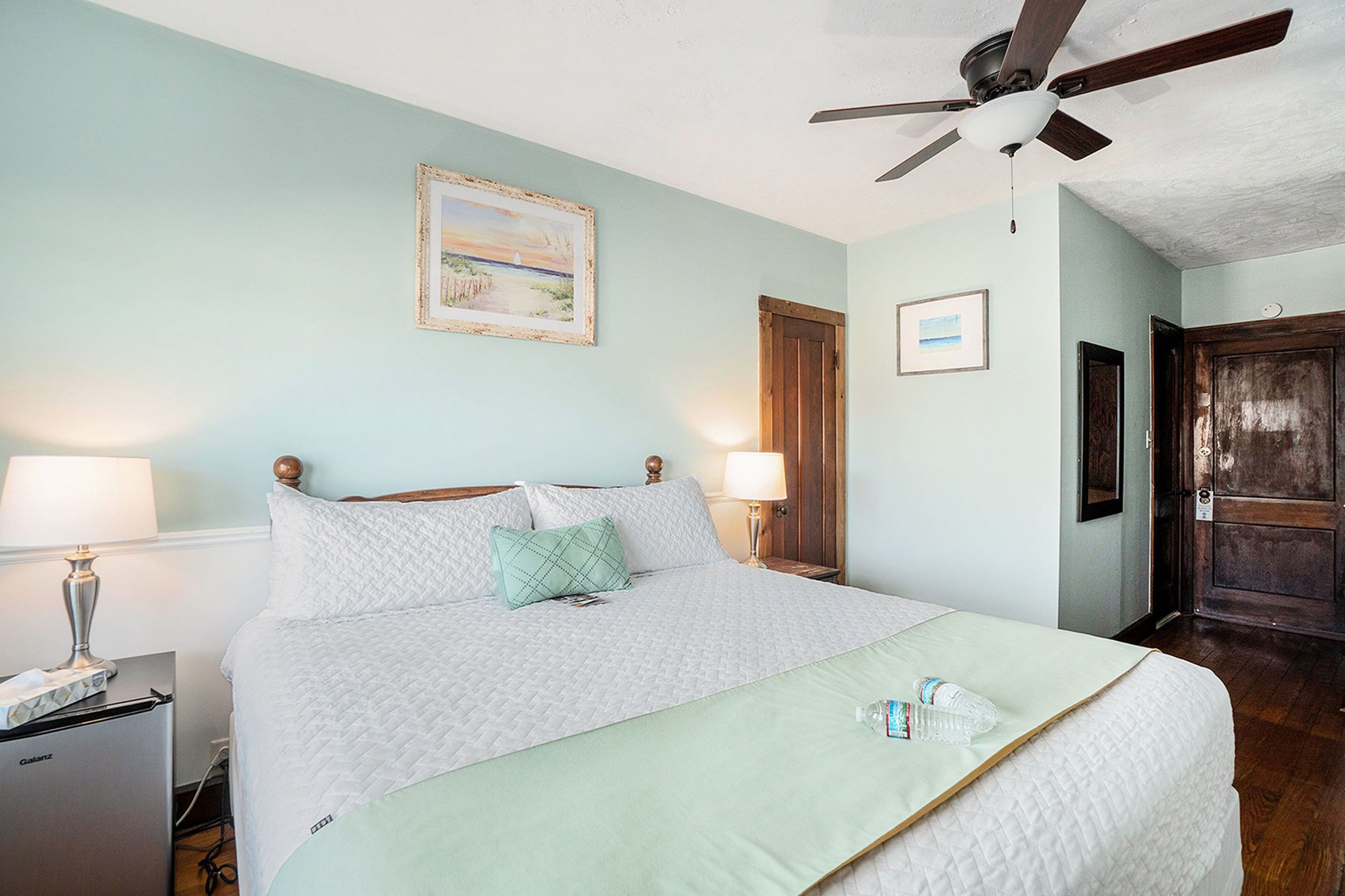 A neatly made bed with a quilted white duvet, a green pillow, and a light green throw blanket. There is a ceiling fan above the bed, a bedside lamp, and a small table with a lamp and a bottle.