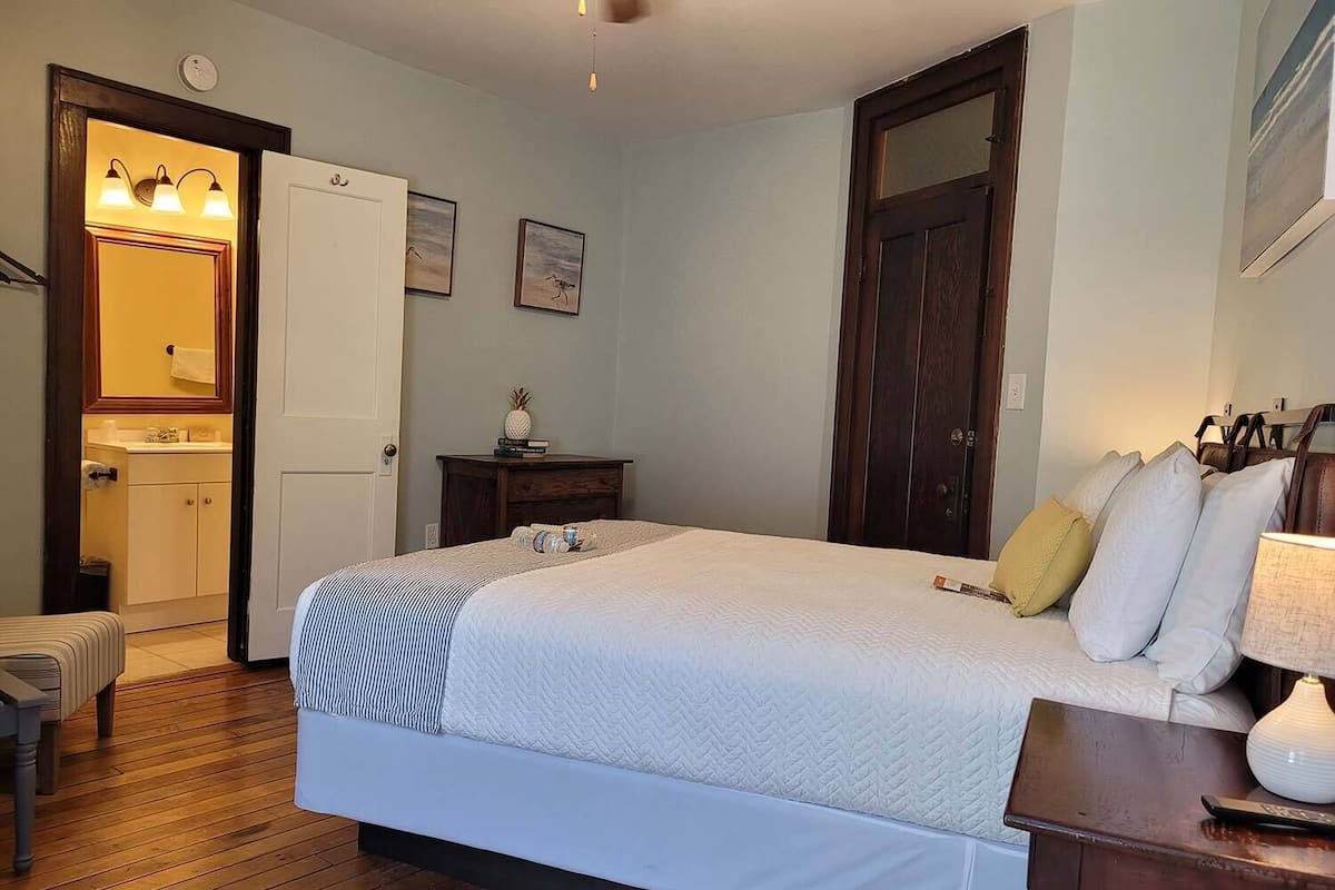 Un dormitorio con una cama grande, una mesita de noche con lámpara, suelo de madera, una puerta y una pequeña mesita de noche con libros y una piña.