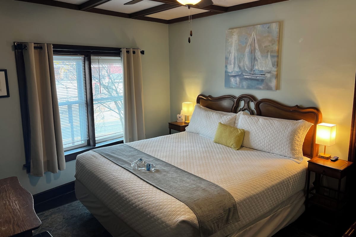 Una habitación de hotel con una cama grande, dos lámparas de noche, un ventilador de techo, una ventana con cortinas y un cuadro en la pared.