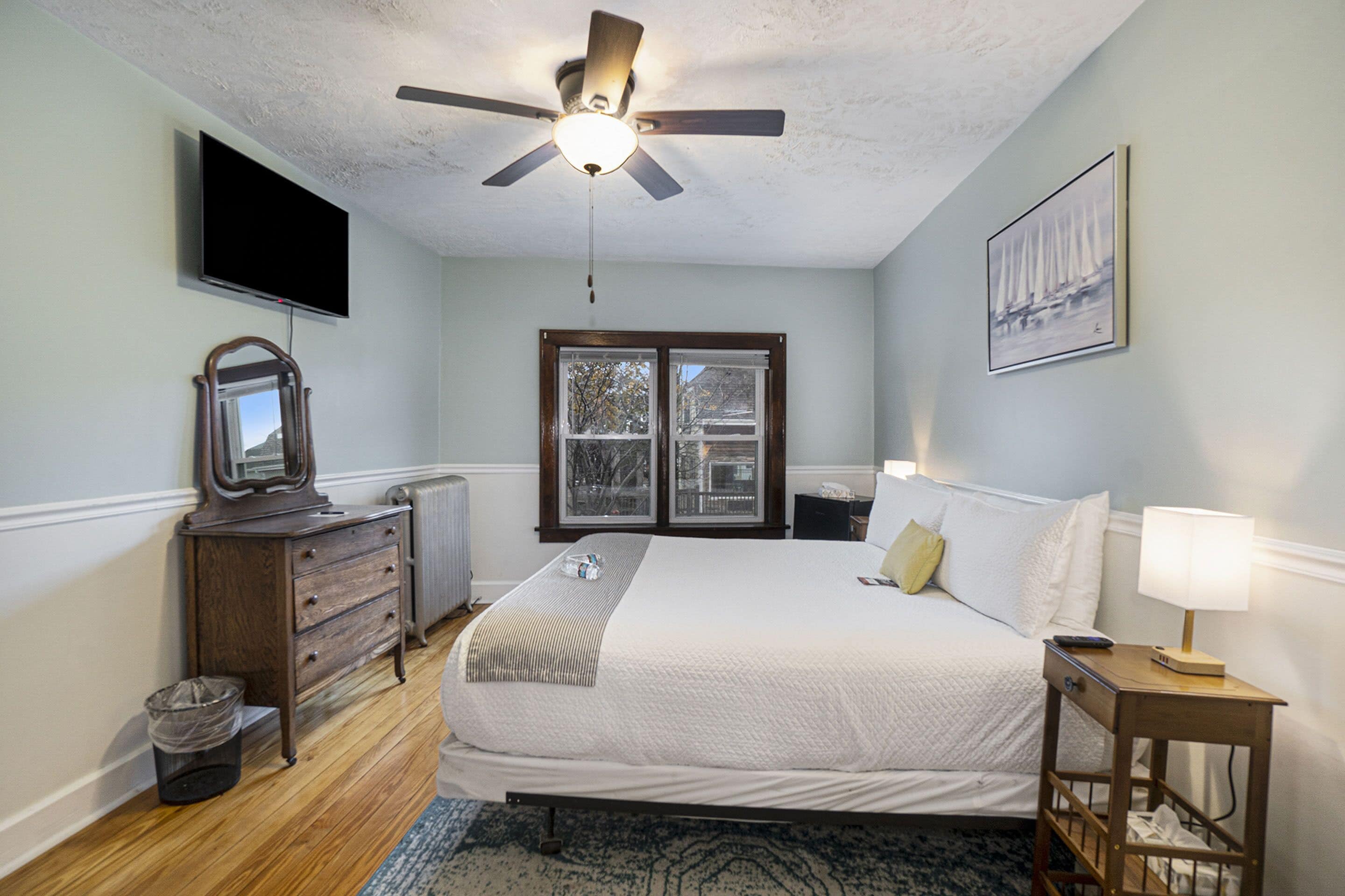 Une chambre à coucher avec un lit, un ventilateur de plafond, une commode, un téléviseur, une fenêtre et un tableau au mur.