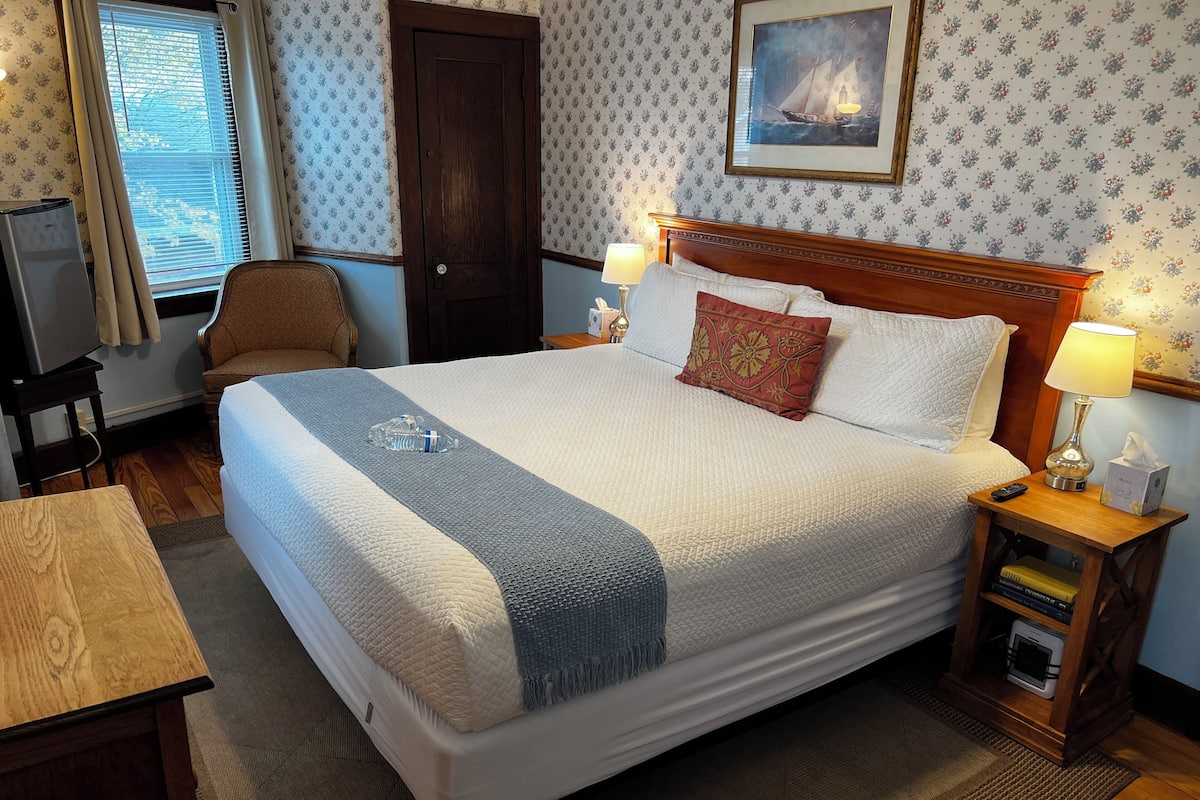 Una cama bien hecha con cabecera de madera, una mesita de noche con lámpara y un cuadro en la pared.
