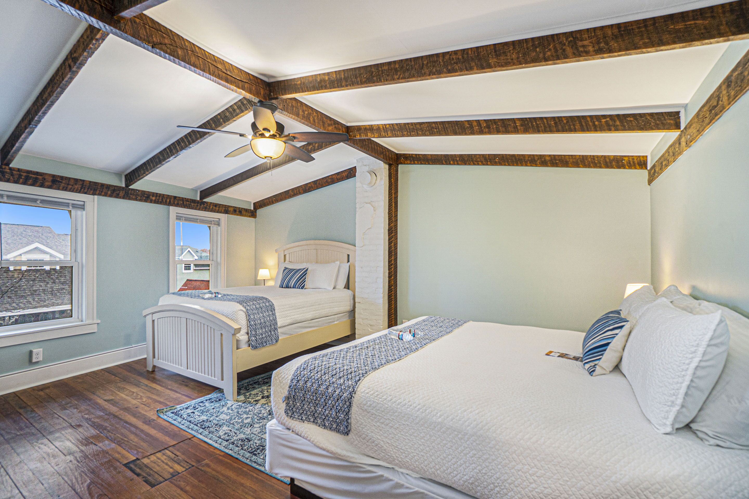 A bedroom with two beds, wooden beams, a ceiling fan, and a view of a house through a window.