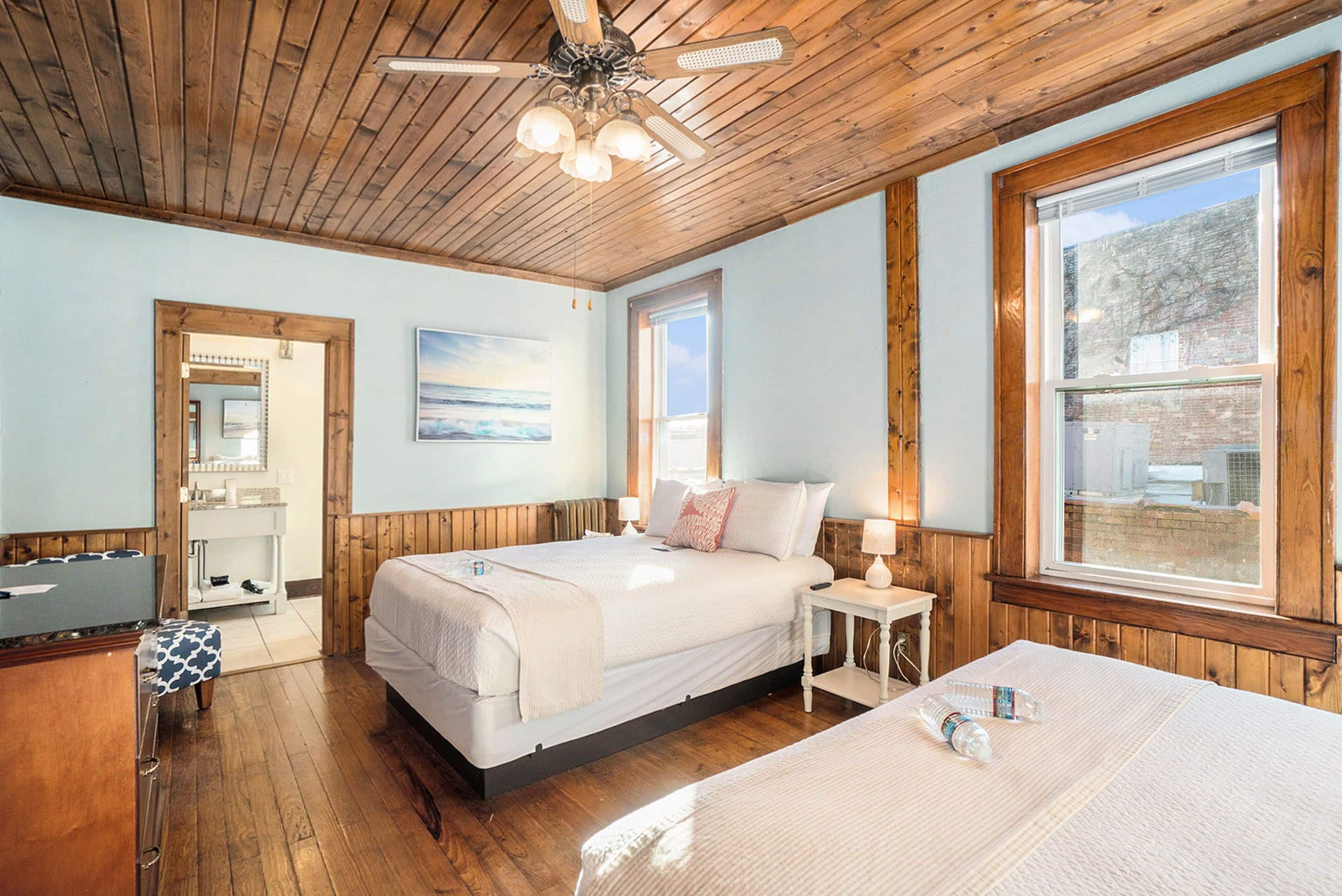 Une chambre avec un plafond en bois, une grande fenêtre, un lit avec une literie blanche, une table de chevet avec une lampe et un plancher en bois.