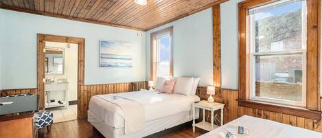 Une chambre avec un plafond en bois, une grande fenêtre, un lit avec une literie blanche, une table de chevet avec une lampe et un plancher en bois.