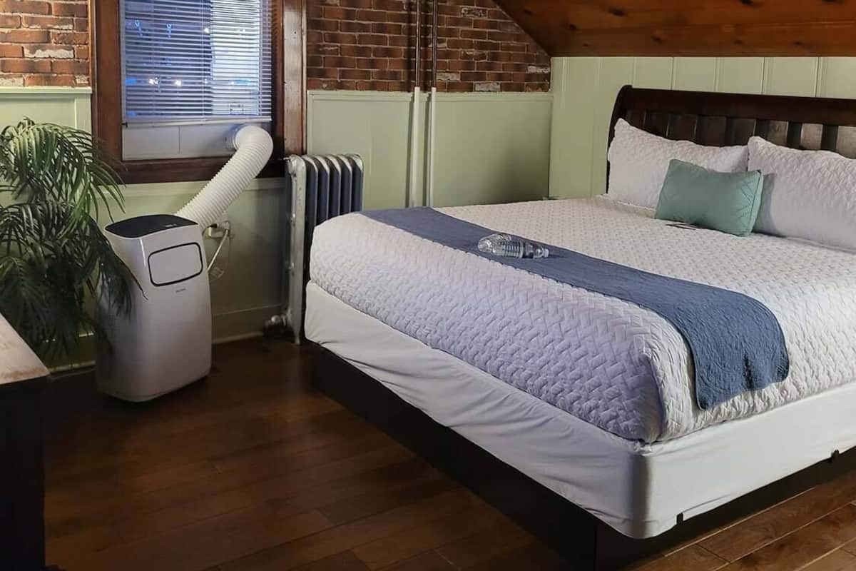 Un dormitorio con techo inclinado, ventilador de techo, cama con colcha blanca y azul, radiador y ventana con persianas.