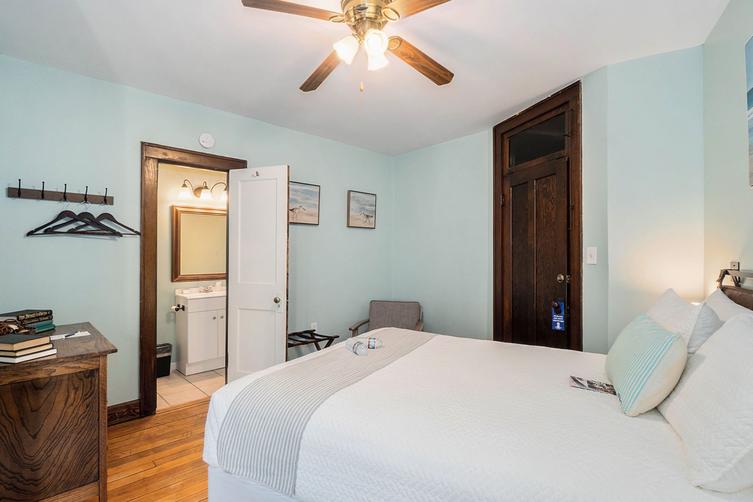 A bedroom with a bed, a ceiling fan, a wooden dresser, a chair, and a door.