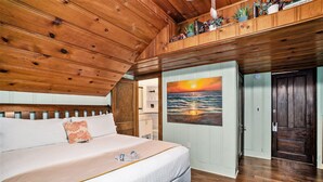 Quarto com teto de madeira, cama com edredom branco, pintura de um pôr do sol e porta de madeira.