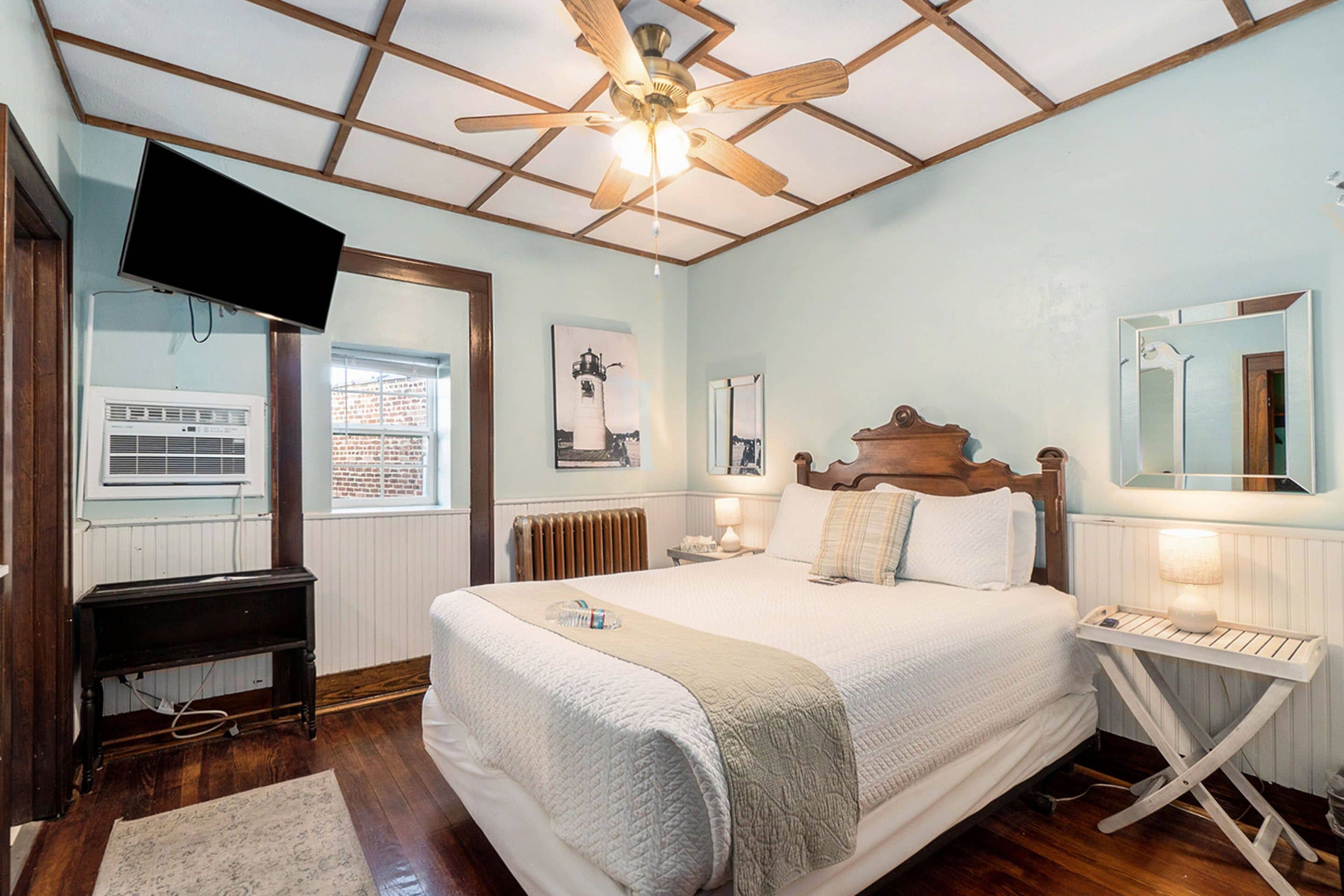 Une chambre avec un plafond en bois, un lit avec une literie blanche, une table de chevet avec une lampe, une commode, un téléviseur fixé au mur et une fenêtre donnant sur un bâtiment en briques.