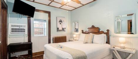Une chambre avec un plafond en bois, un lit avec une literie blanche, une table de chevet avec une lampe, une commode, un téléviseur fixé au mur et une fenêtre donnant sur un bâtiment en briques.