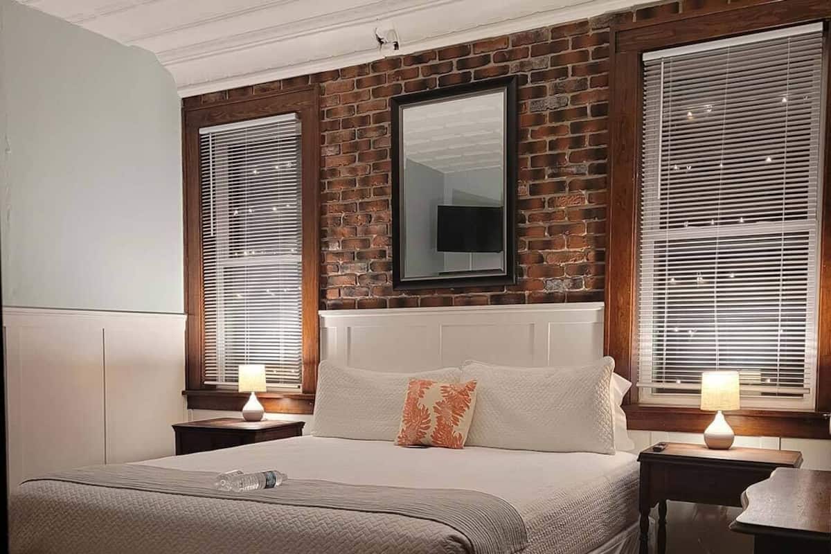 Un dormitorio con pared de ladrillo, una cama grande con cabecera, dos mesitas de noche con lámparas y una ventana con persianas.