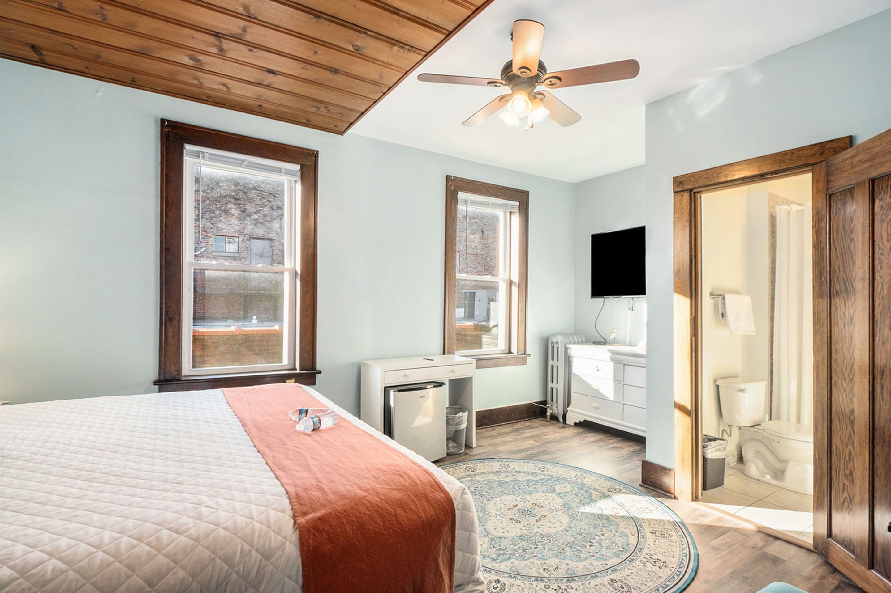 A bedroom with a bed, a desk, a refrigerator, a window, a ceiling fan, and a bathroom with a toilet and sink.