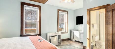 A bedroom with a bed, a desk, a refrigerator, a window, a ceiling fan, and a bathroom with a toilet and sink.