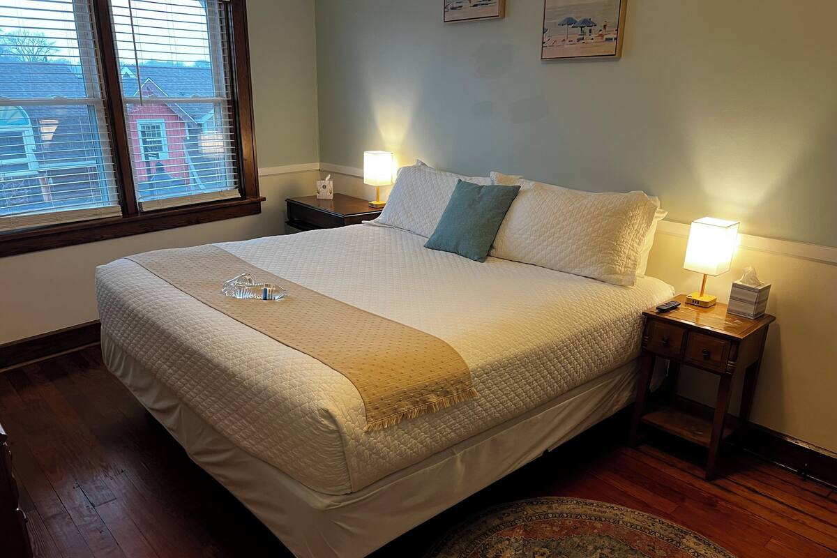 Una cama bien tendida con colcha, dos almohadas, una lámpara de noche y una mesita de noche con un dispensador de pañuelos y un control remoto.