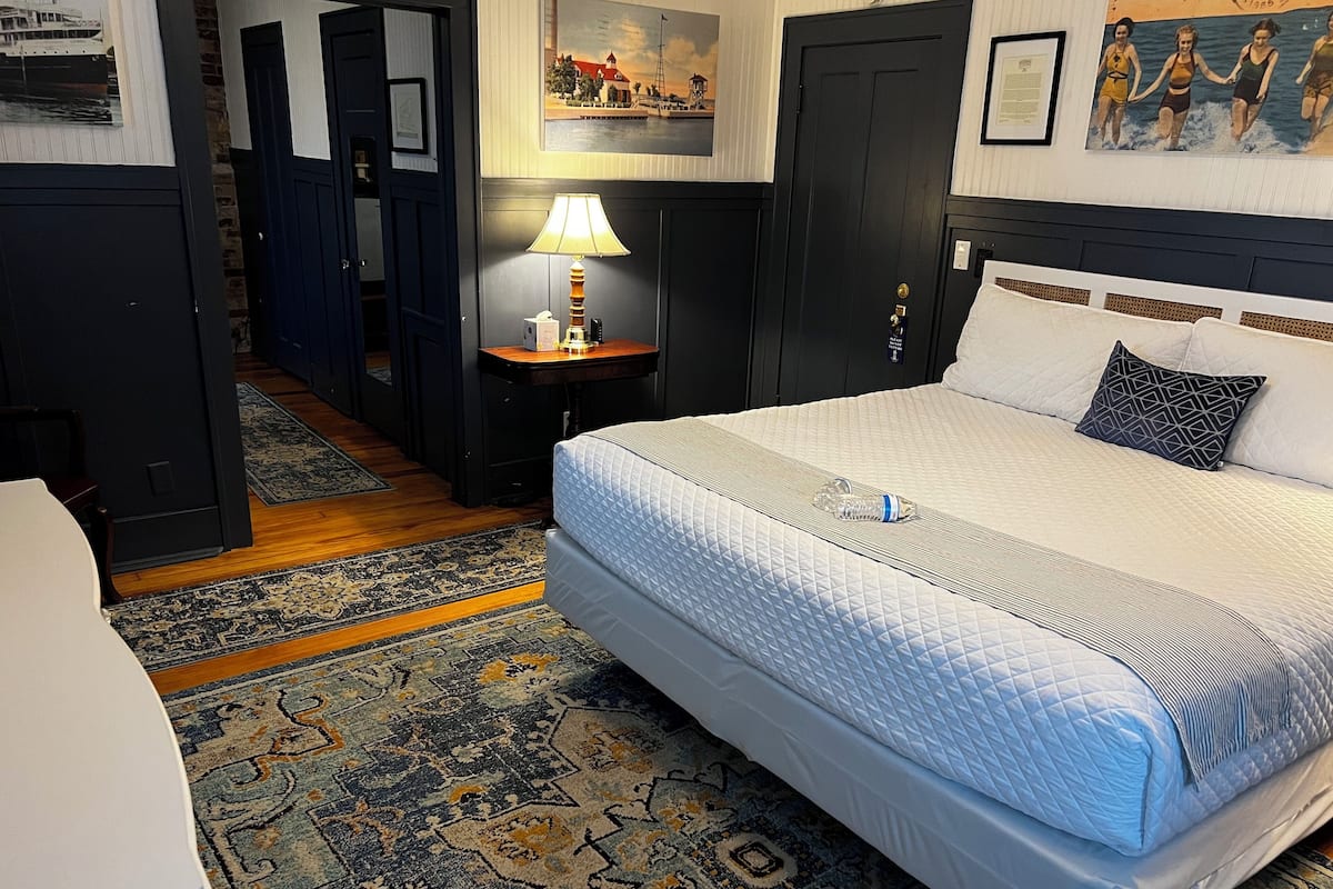 Una habitación de hotel con una cama grande, una mesita de noche con lámpara, una alfombra estampada y un cuadro en la pared.