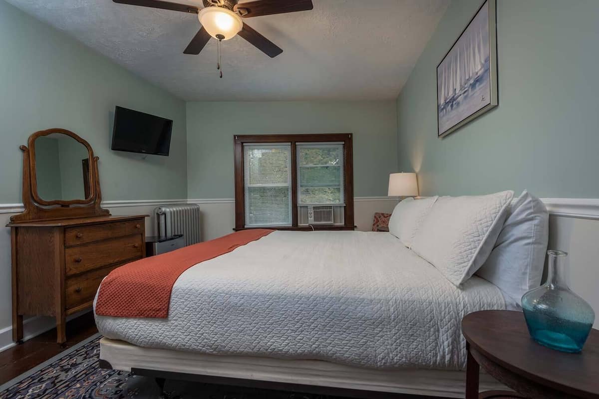 Un dormitorio con cama, cómoda, televisor, ventana con persianas, ventilador de techo y un cuadro en la pared.