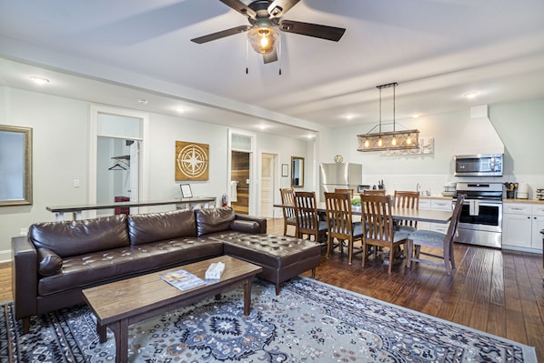 A spacious living room with a leather sectional sofa, a coffee table, and a patterned rug. There is a dining area with wooden chairs and a chandelier. A kitchen with stainless steel appliances is visible in the background. - Historic Hotel Nichols (South Haven)