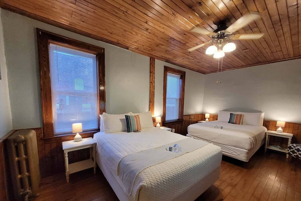 Una habitación con dos camas, pisos de madera, un ventilador de techo y una ventana con persianas.