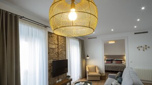 A modern living room with a large flat-screen TV, a wooden bench, a round glass coffee table, a sofa with patterned cushions, and a unique spherical hanging light fixture.