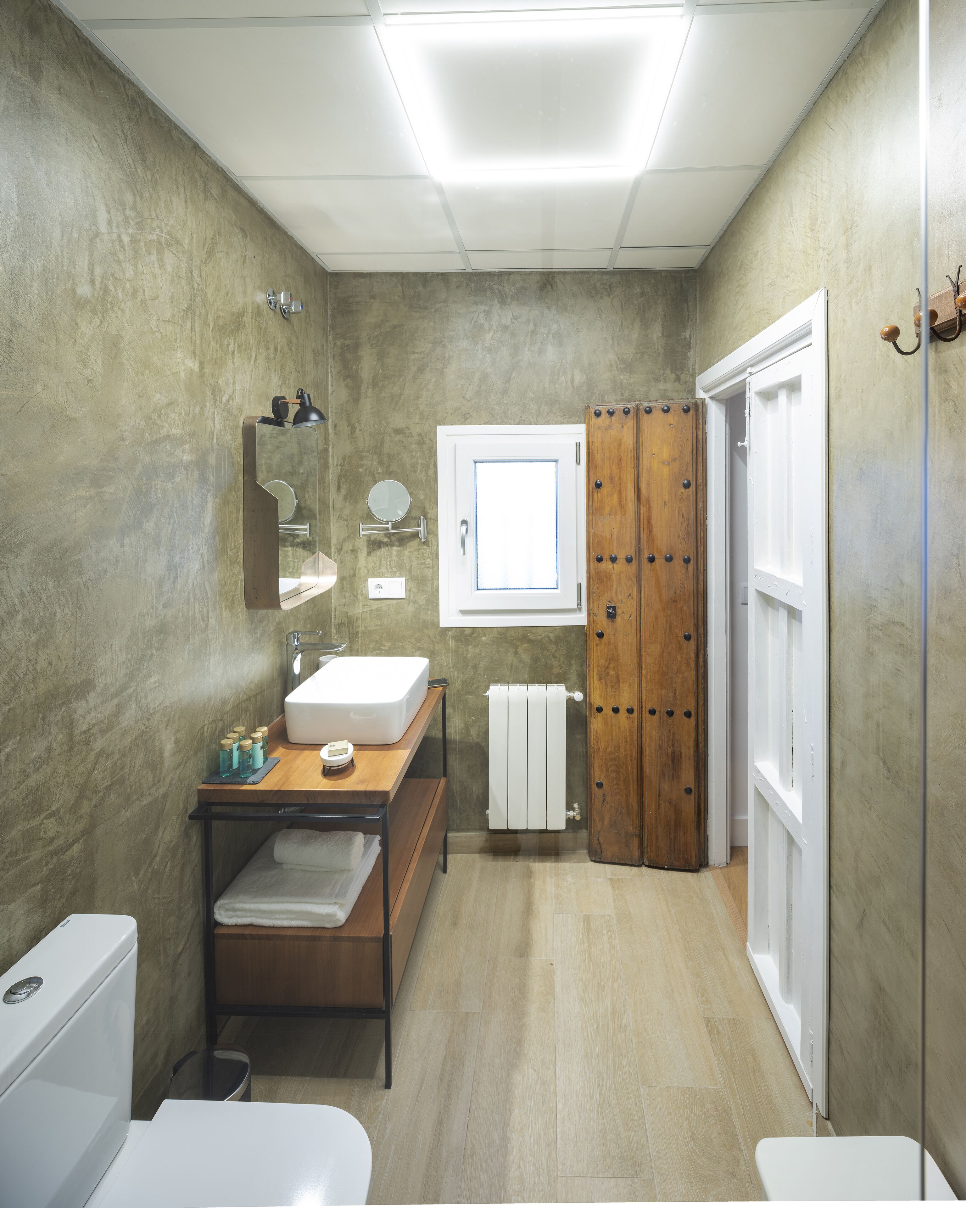 Un baño con tocador de madera, lavamanos blanco, espejo, perchero para toallas y puerta de madera.