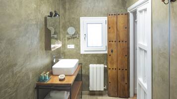 Un baño con tocador de madera, lavamanos blanco, espejo, perchero para toallas y puerta de madera.