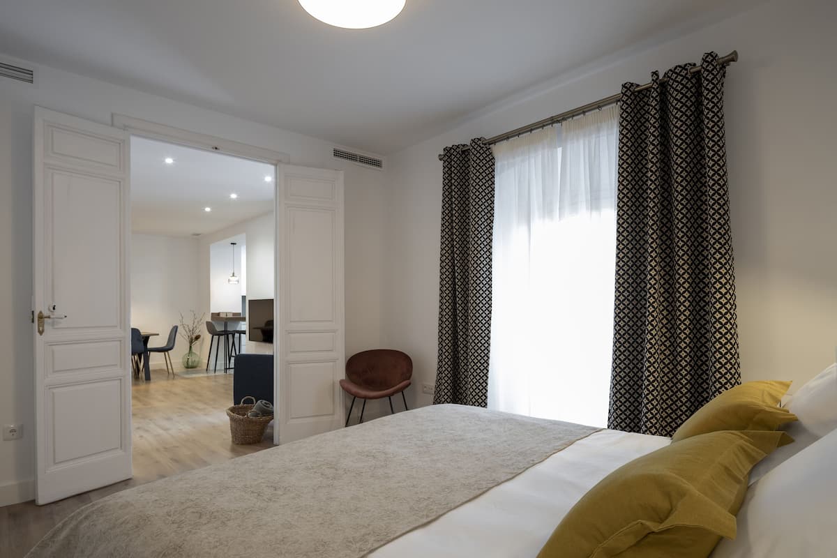 A bedroom with a bed, a chair, a bedside table, and a window with patterned curtains.