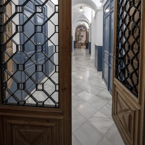 A hallway with a geometric patterned door, marble flooring, and a view into a room with a large window and a decorative ceiling.