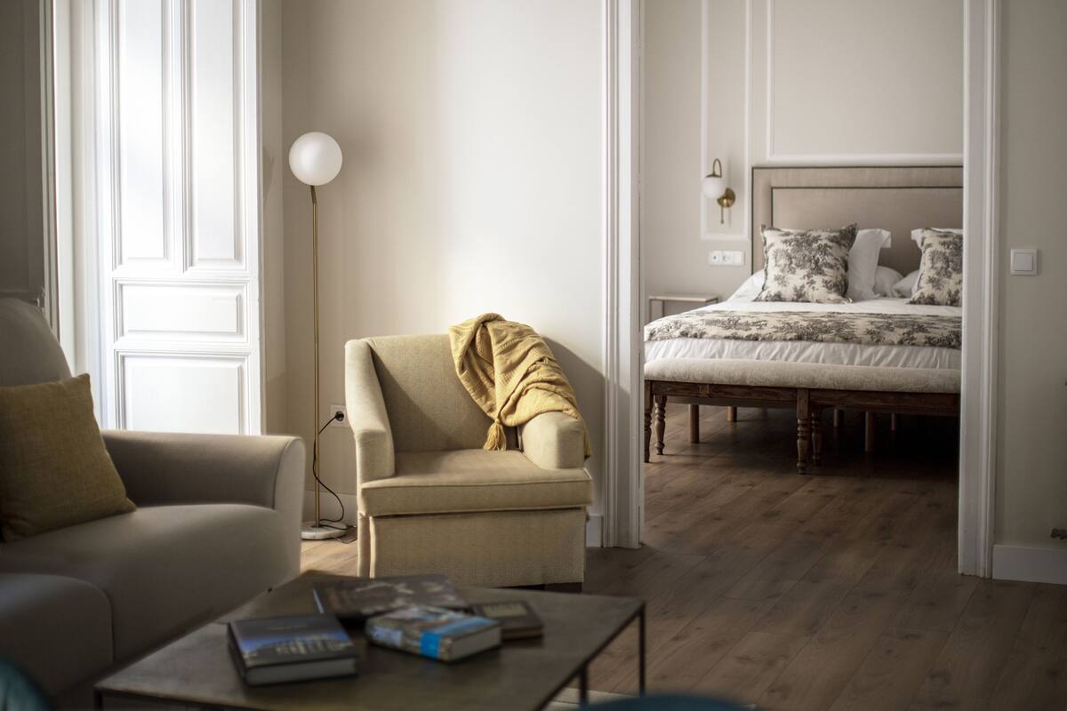 A modern living room with a sofa, armchair, and coffee table. A bed is visible in the background.