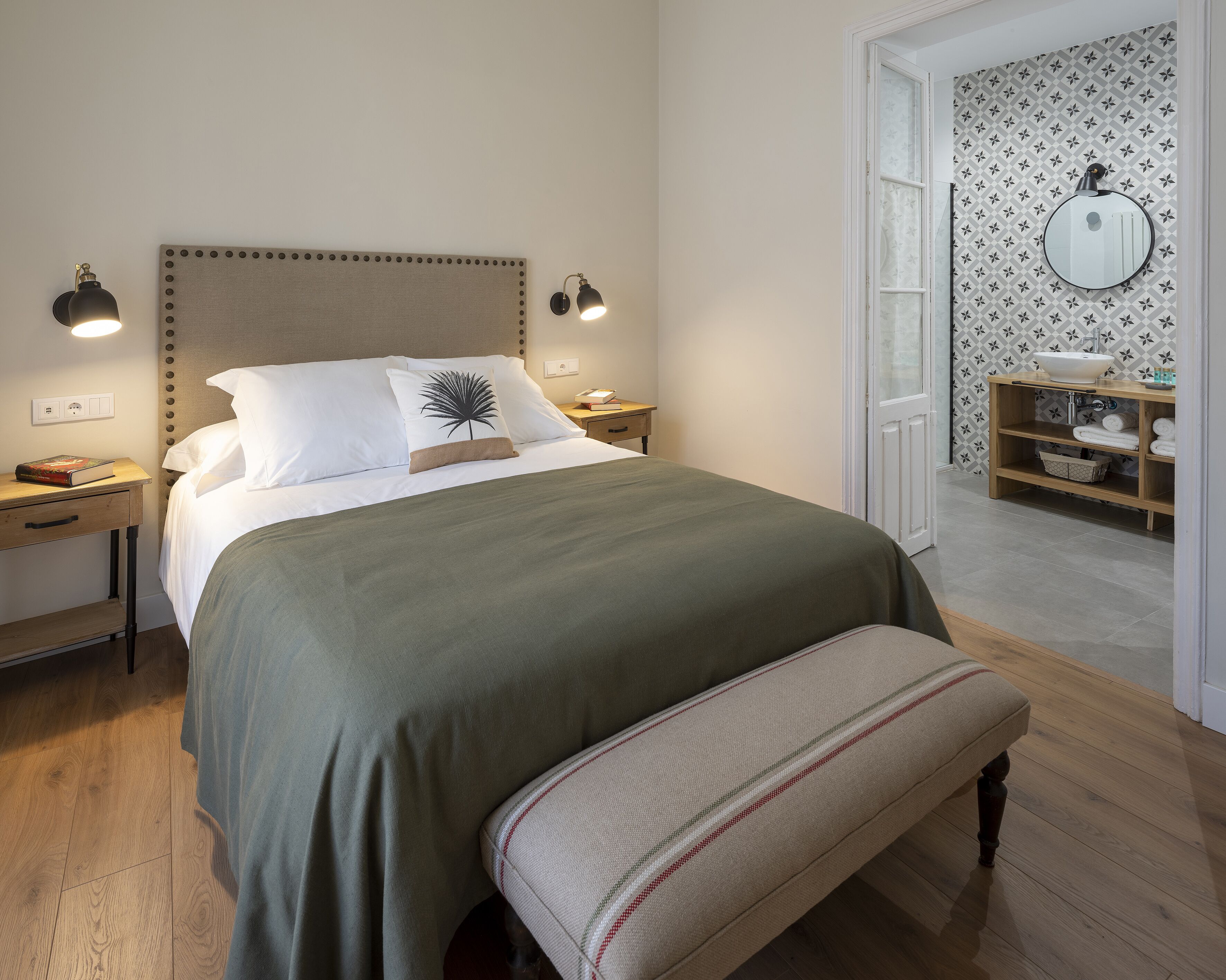 A neatly made bed with a green bedspread and a single decorative pillow, flanked by two bedside lamps. A wooden nightstand with a book and a small drawer is to the left. The room features a wooden floor and a bench at the foot of the bed.