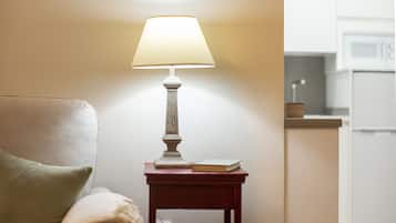 Una lámpara sobre una mesita de noche junto a un libro, una silla con un cojín y una cocina al fondo.