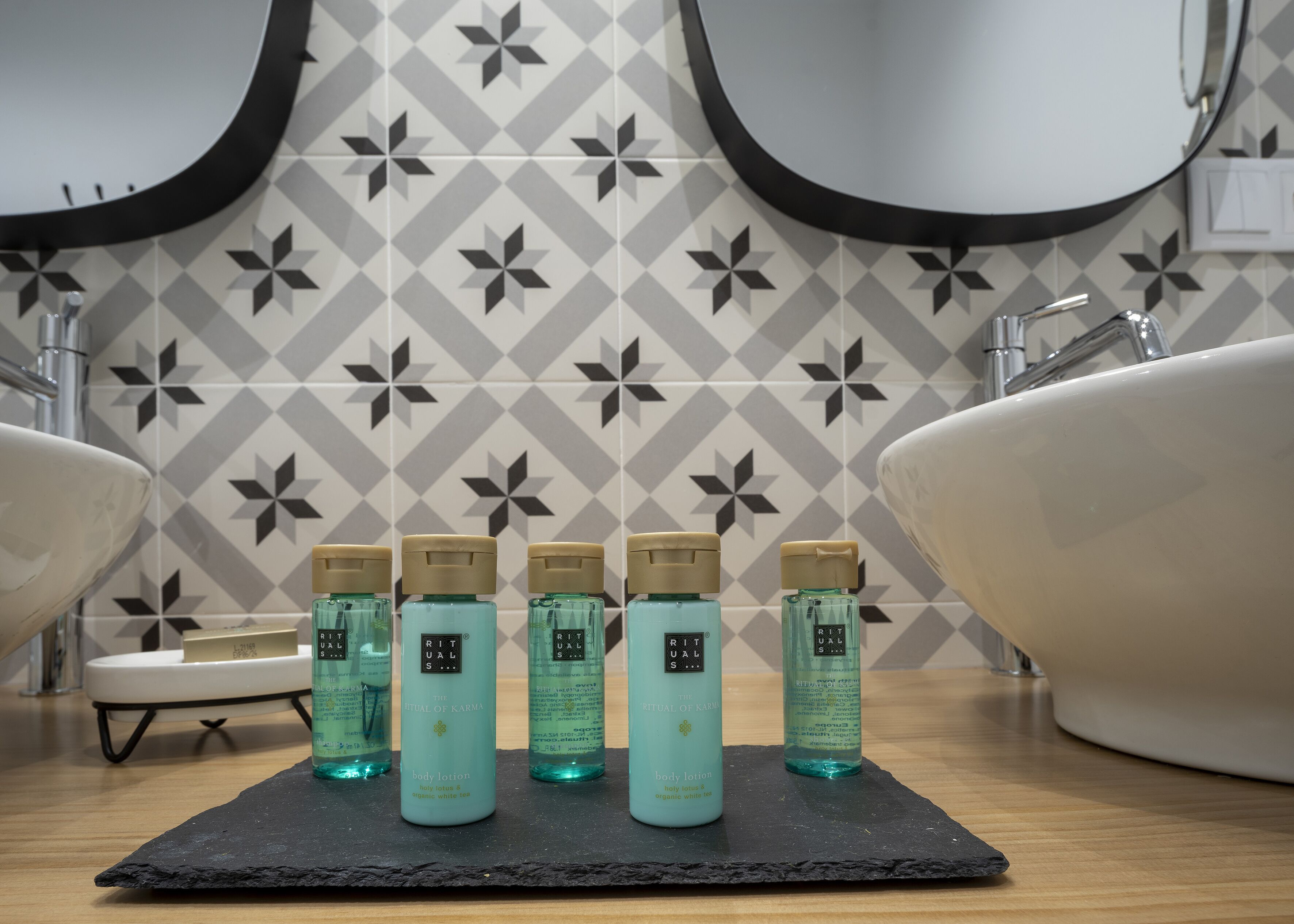 A bathroom with a geometric patterned wall, two sinks, and four bottles of body lotion on a black slate.